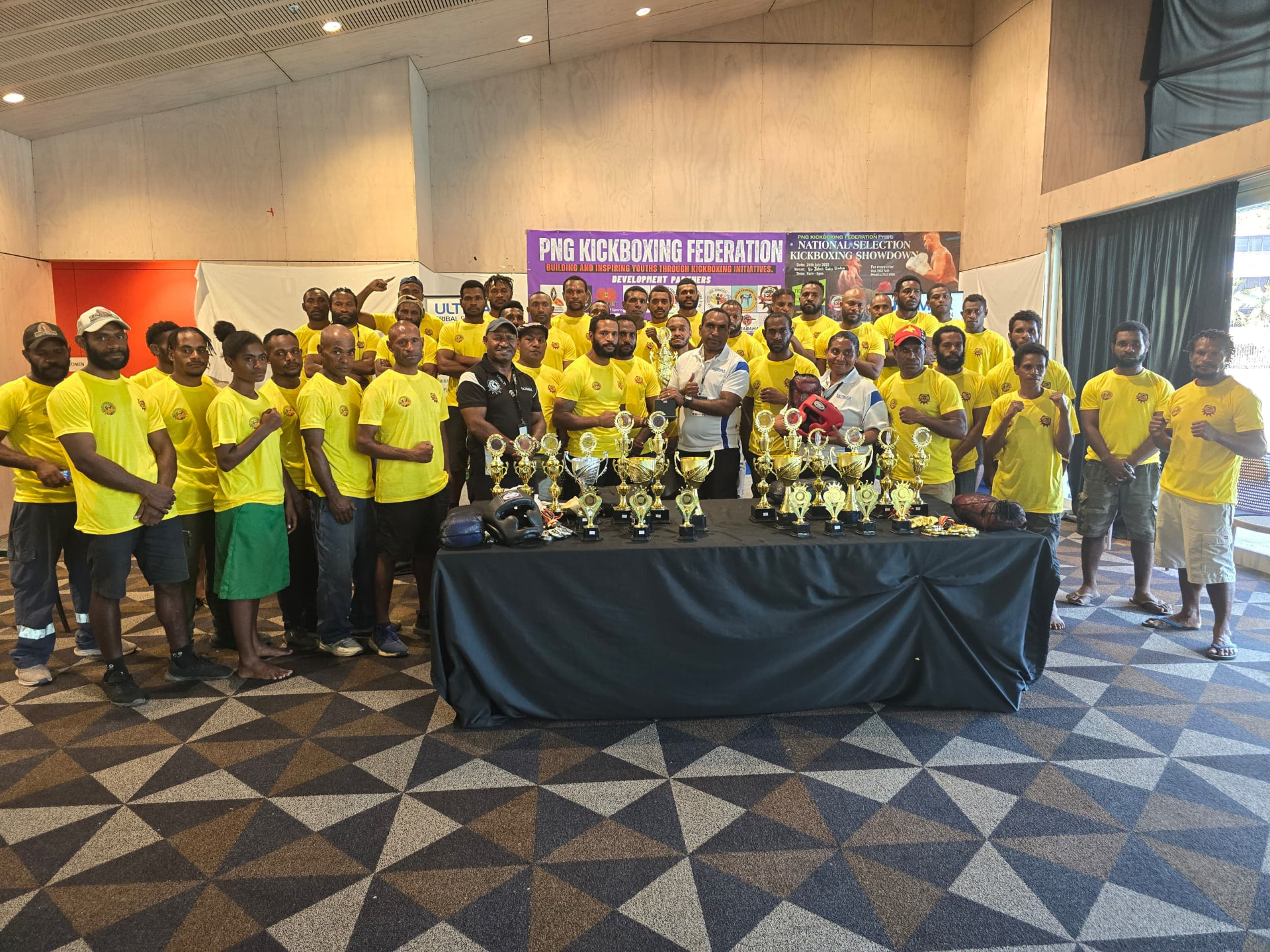 PNG Kickboxing Federation athletes in yellow shirts gathered behind a table of championship trophies